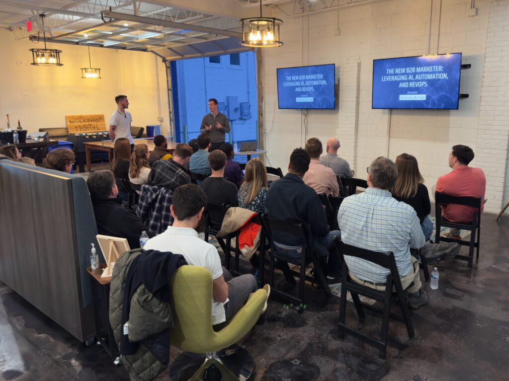 A seated crowed watching one person introduce another person before the present slides on a tv screen mounted to the wall.