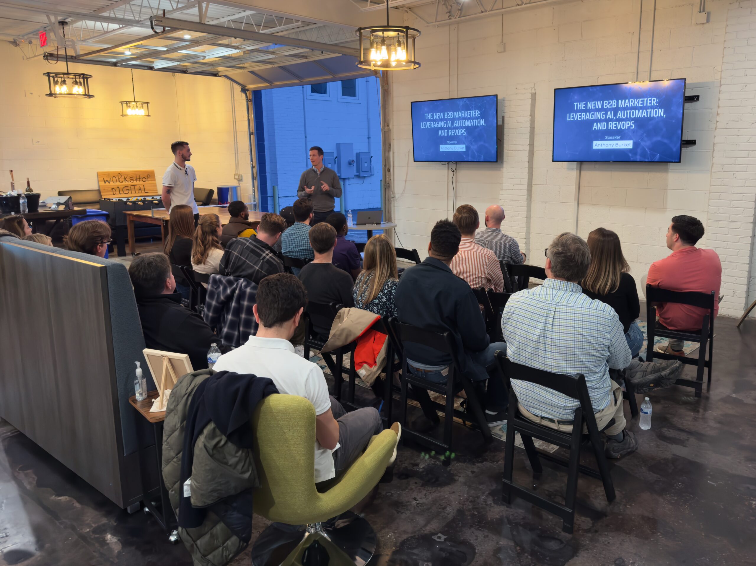 A seated crowed watching one person introduce another person before the present slides on a tv screen mounted to the wall.
