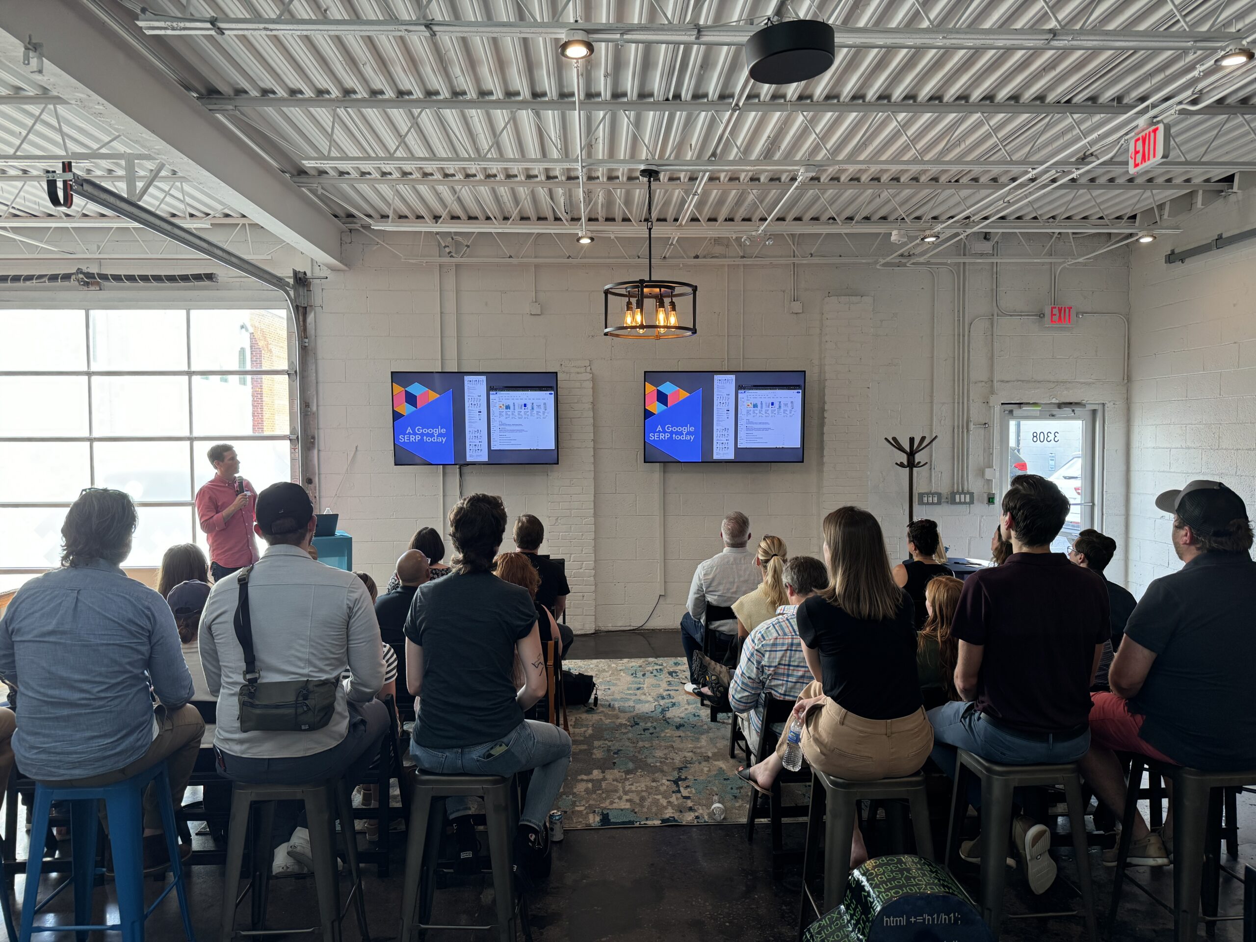 A speaker presents slides displayed on two monitors attached to the wall in front of an audience seated in a modern, industrial-style room with white walls and a high ceiling.
